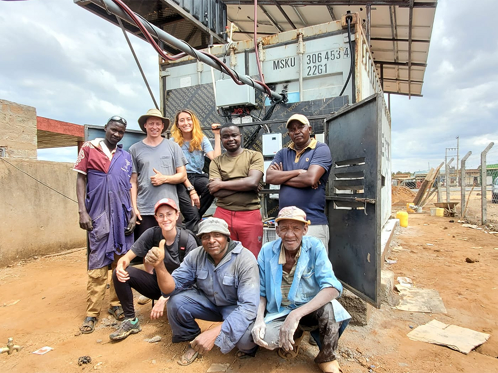 A diverse group of 8 people, five standing and three crouching, pose in front of a shipping container that has been modified for use for cold storage.