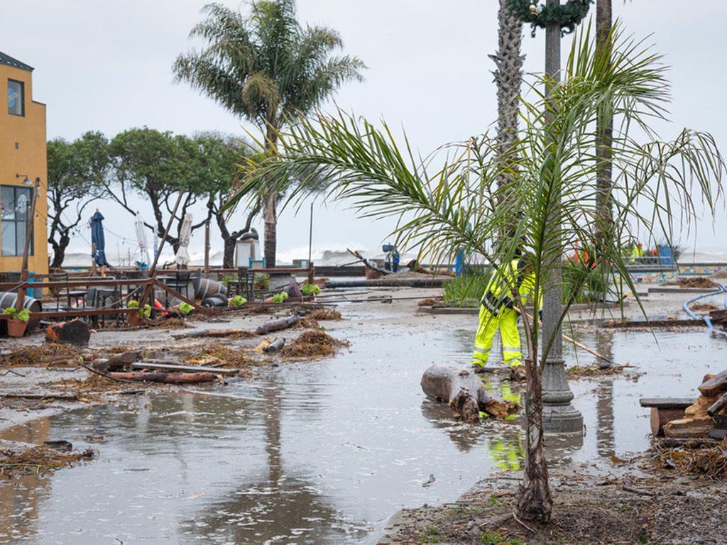 A worker cleans up a flooded, debris-filled street after a bomb cyclone hit Santa Cruz, CA.