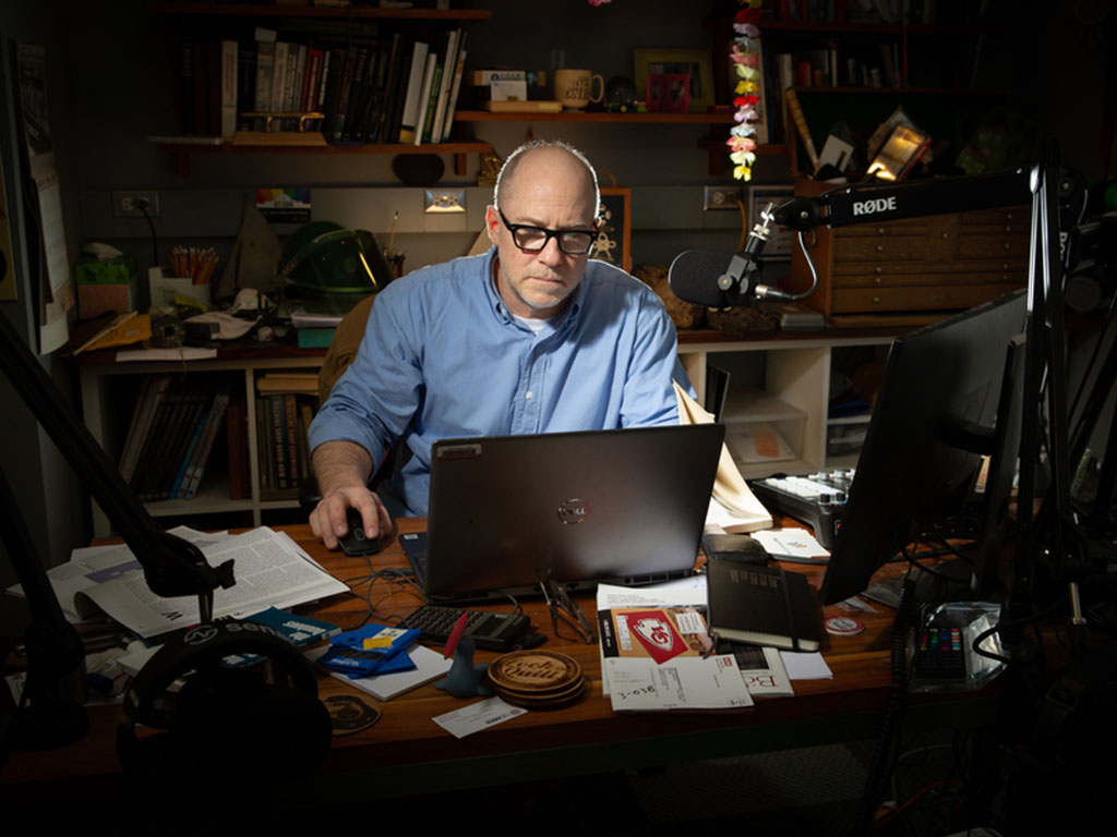 Danny Braunstein, sitting in a pool of light in an otherwise-dim room, looks at a laptop. There is a professional-grade boom microphone in front of him.