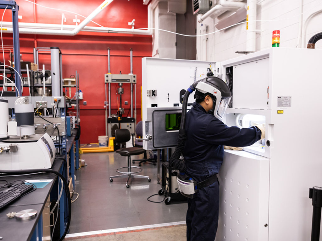 A student wearing protective gear including a full helmet and face shield with air supply works at a refrigerator-sized machine