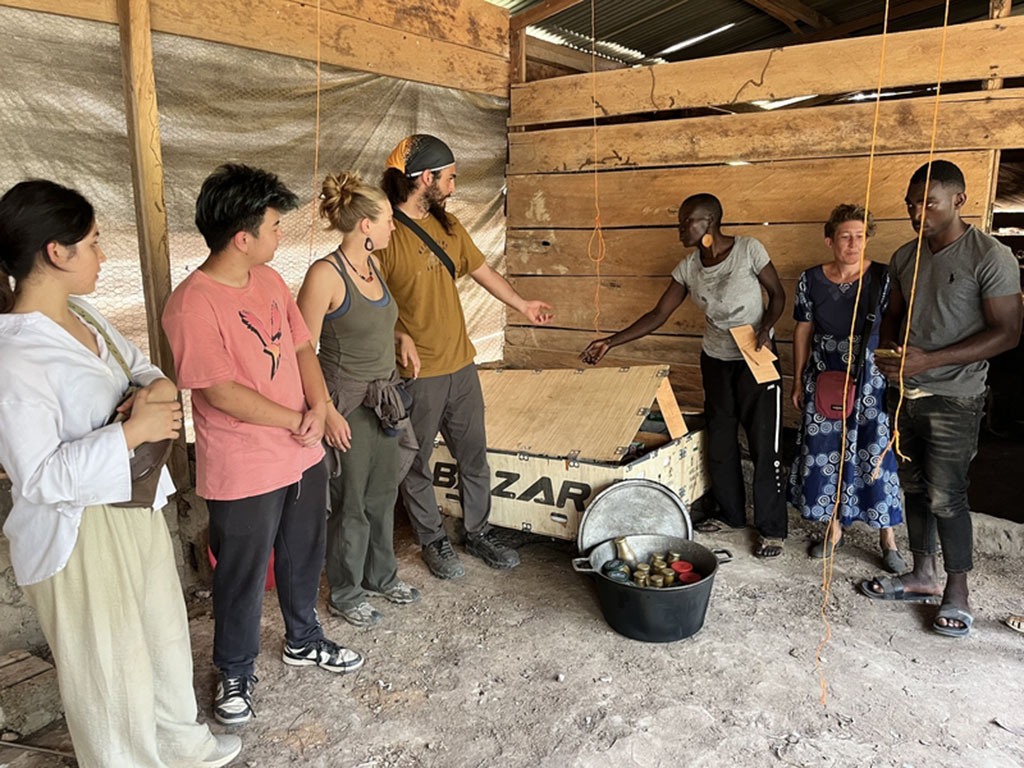 Seven people standing by a wooden box and a load of beeswax thermal batteries in a rough structure with a dirt floor