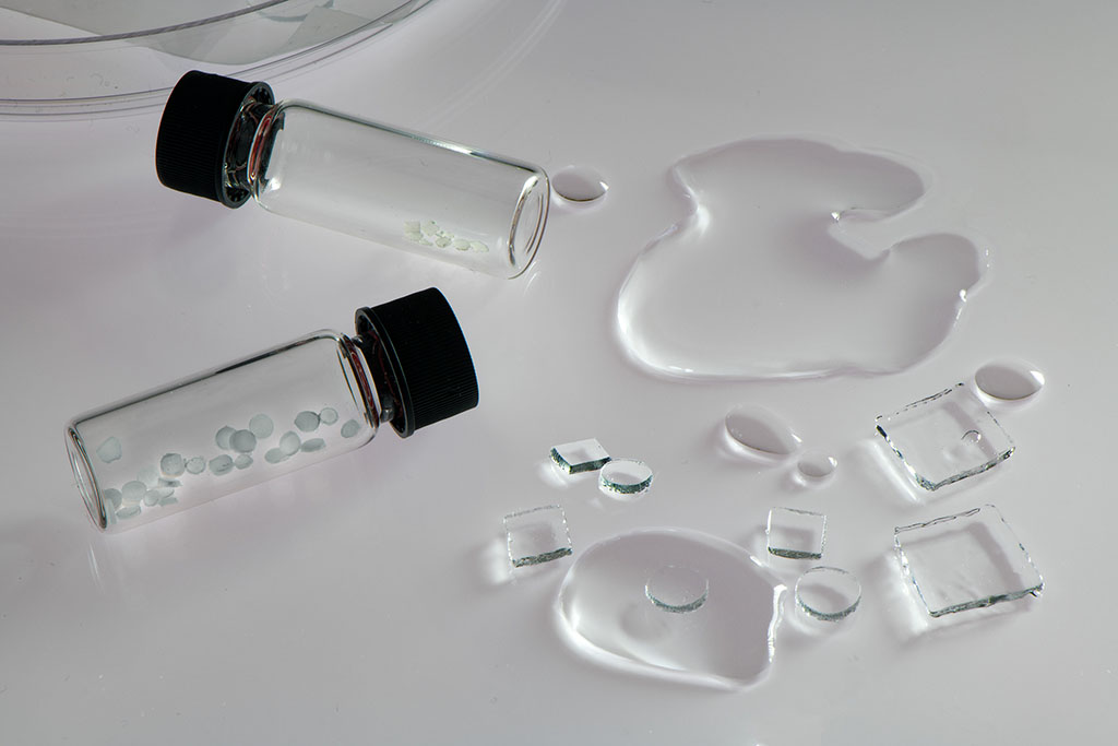 On a white table, 2 small glass vials contain pebble-like translucent chips. Some puddles of water are on the table, as well as circular and square chips of a hydrogel substance.