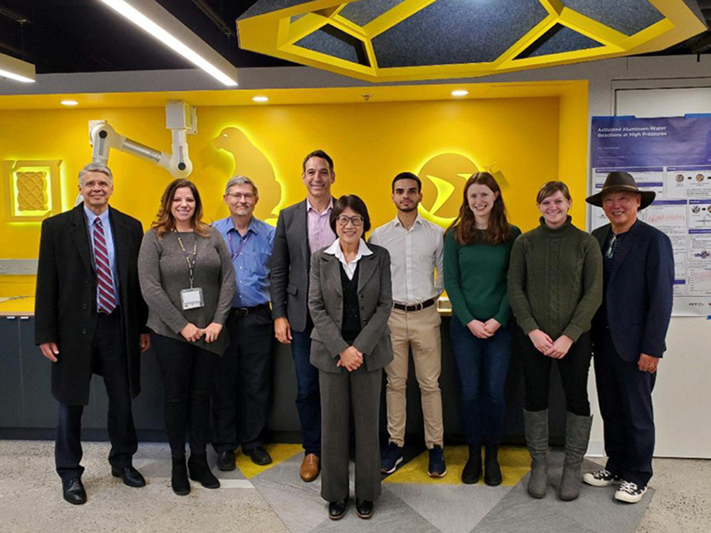 Photo of nine people posing in a line inside a colorful yellow-and-blue workshop