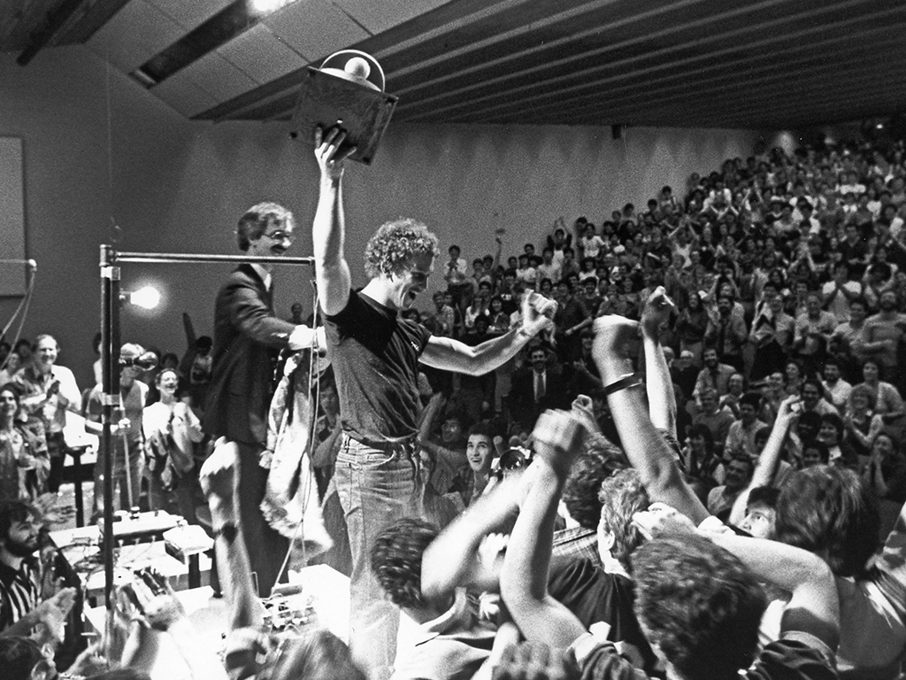 A black and white image of a man holding a trophy, surrounded by students.